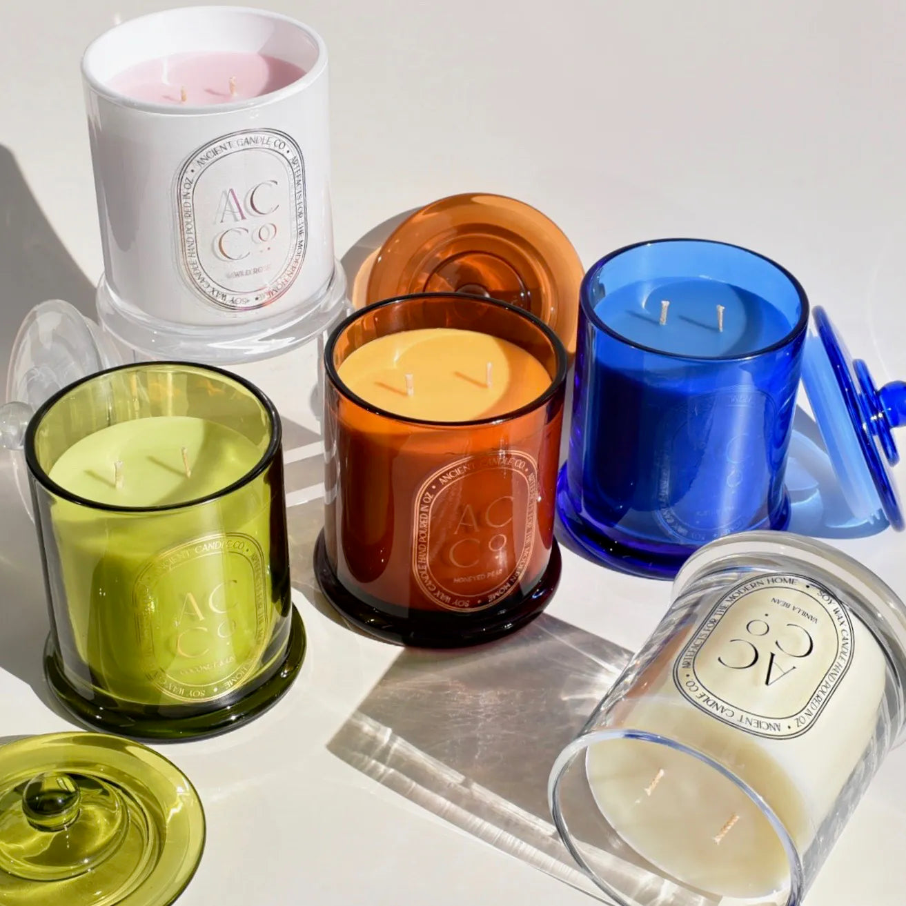 A collection of colorful glass jar candles from Ancient Candle Co, each with a lid. The jars are in various colors: white, orange, blue, green, and two-toned. They are arranged on a white surface with light casting shadows. Each handcrafted candle has a minimalist label design.
