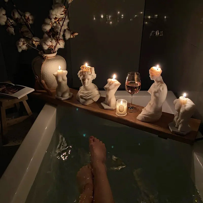 A dimly-lit bathroom scene shows someone's legs stretched out in a bathtub filled with water. A wooden tray across the tub holds five statue-shaped candles with flickering flames, casting a serene candlelight for mental well-being. A glass of wine and cotton stems in a vase sit blissfully in the background.