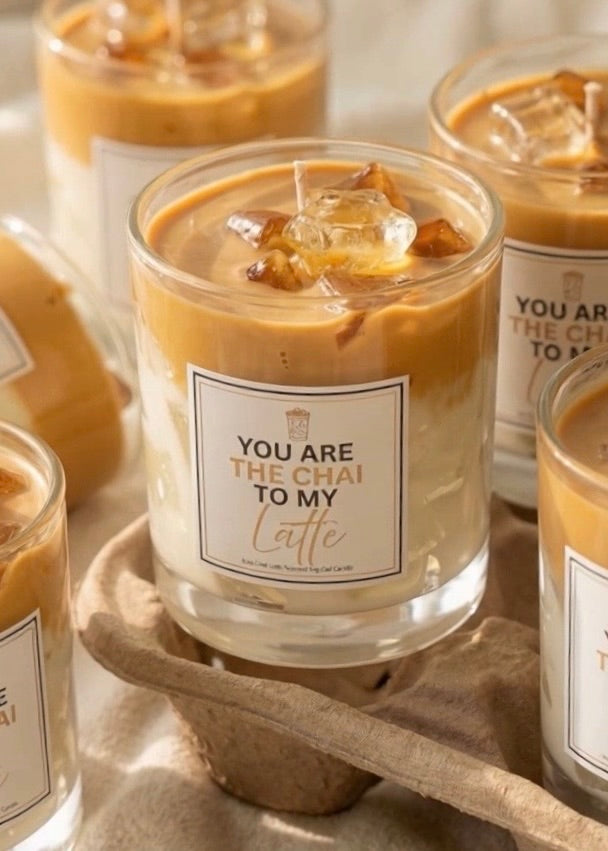 Candle in a glass jar with a label reading 'You are the Chai to my Latte' on a wooden surface.