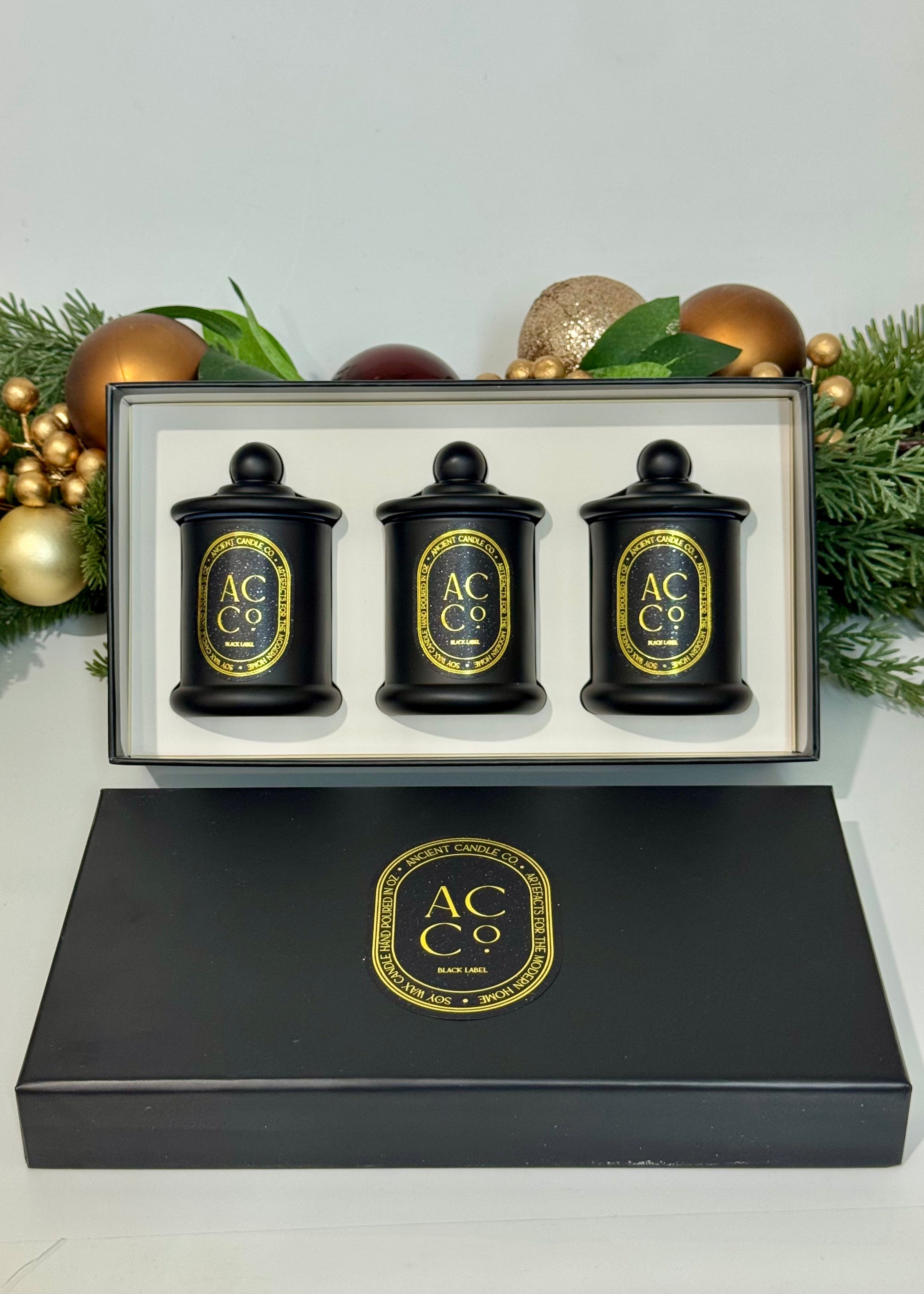 Set of three black candles with gold labels in a decorative box, surrounded by Christmas decorations.