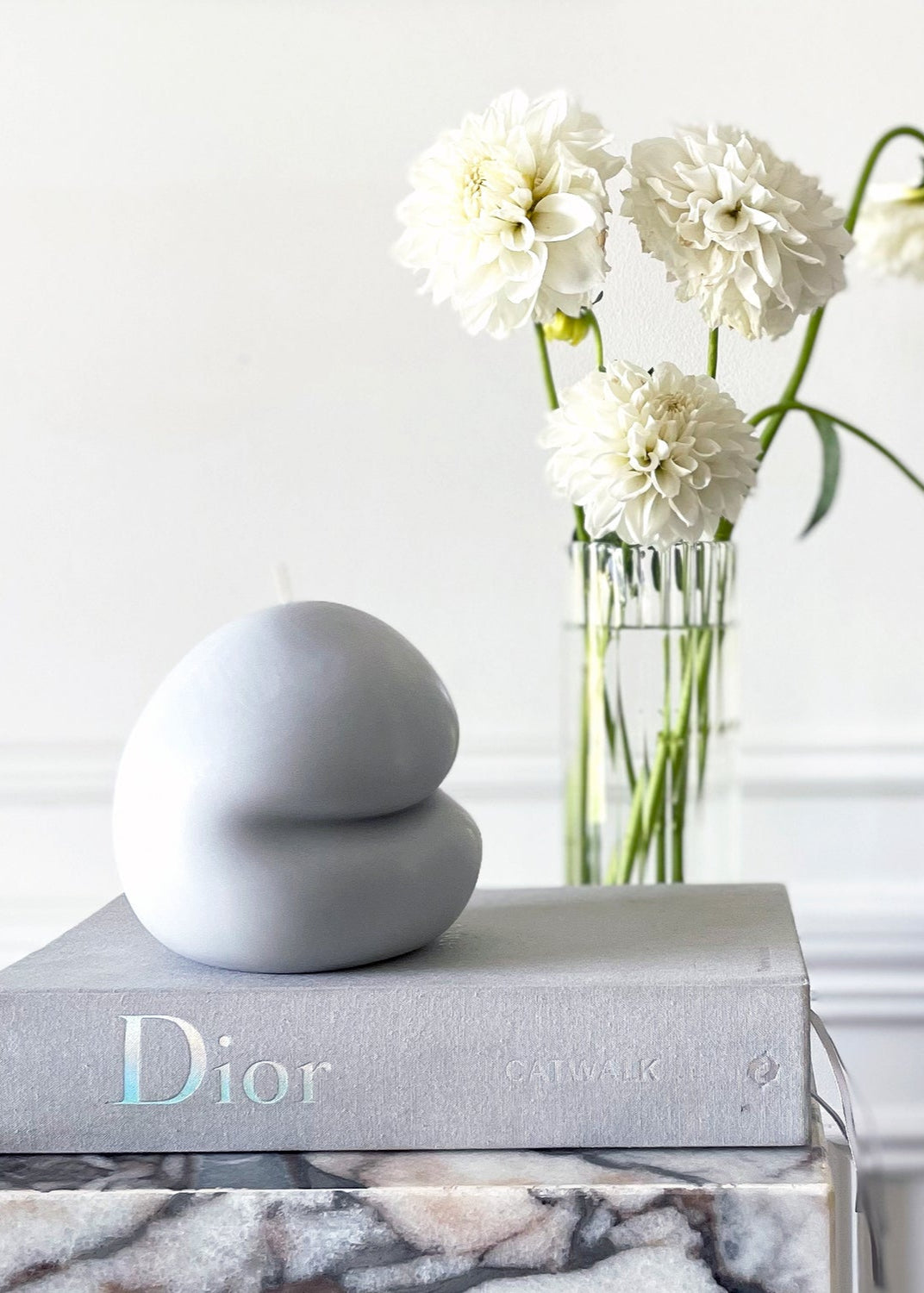 Stack of books with a Dior logo, a decorative blobbie candle and a vase with white flowers on a marble surface.