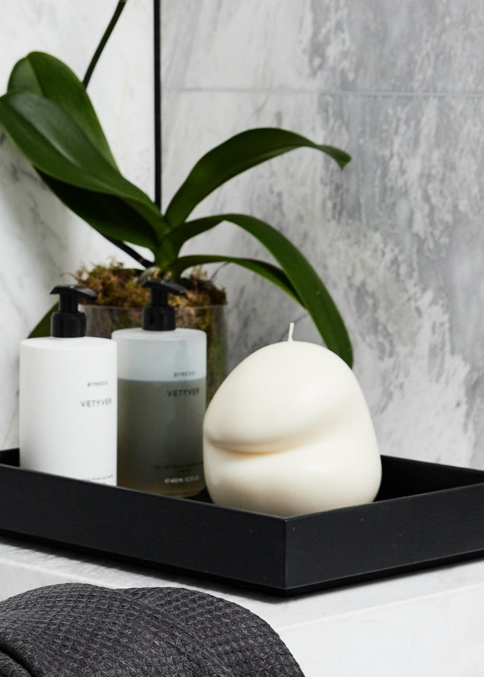 Bathroom setting with black tray holding bottles, a white tulum blobbie candle, and a plant against a gray tiled wall.